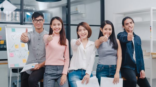 Group of Asia young creative people in smart casual wear smiling and thumbs up in creative office workplace. Diverse Asian male and female stand together at startup. Coworker teamwork concept.